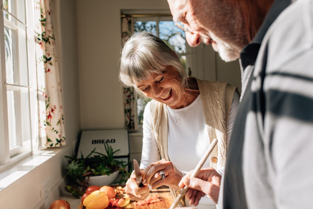 Cheerful woman cutting vegetables while her husband is cooking food.