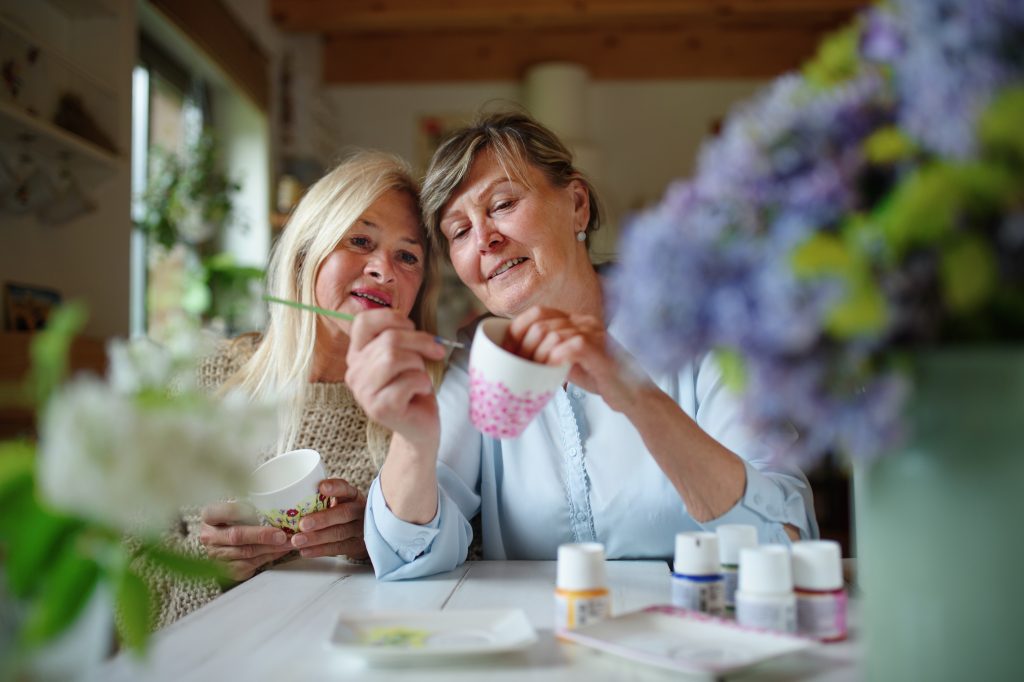 A happy senior woman artist teaching her friend how to paint a ceramic cup.