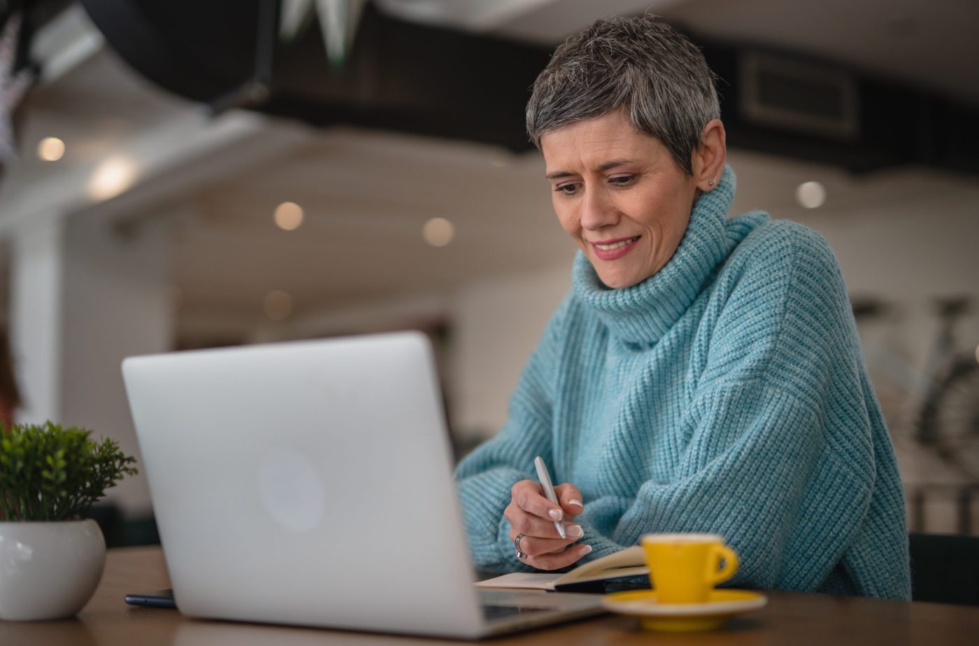 One senior woman in blue sweater sitting, working on laptop and writes in a notebook, modern business senior woman casual concept