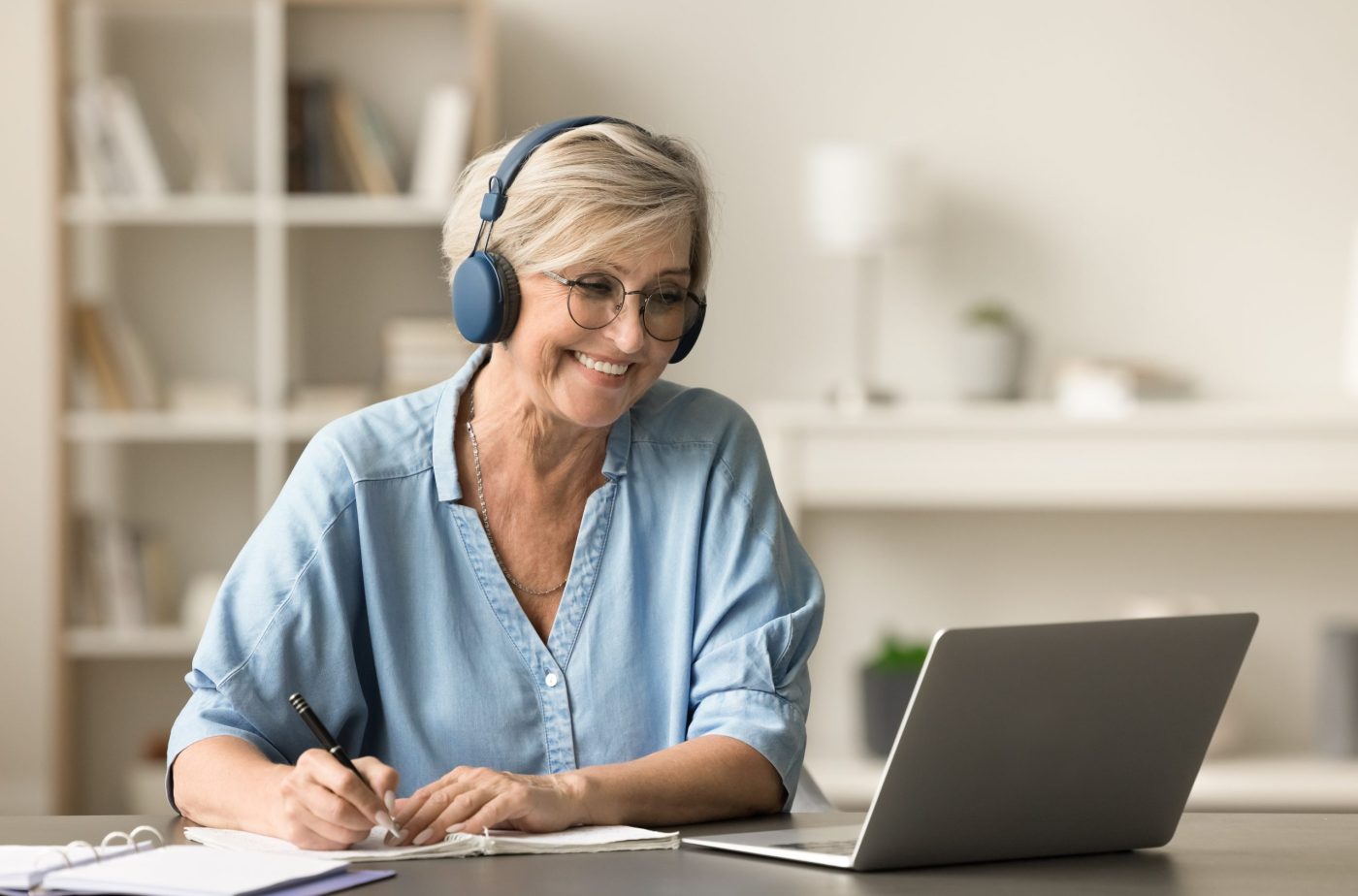 Happy senior retired student woman in earphones and glasses studying on Internet from home, writing notes at laptop, watching online educating lecture, smiling, laughing