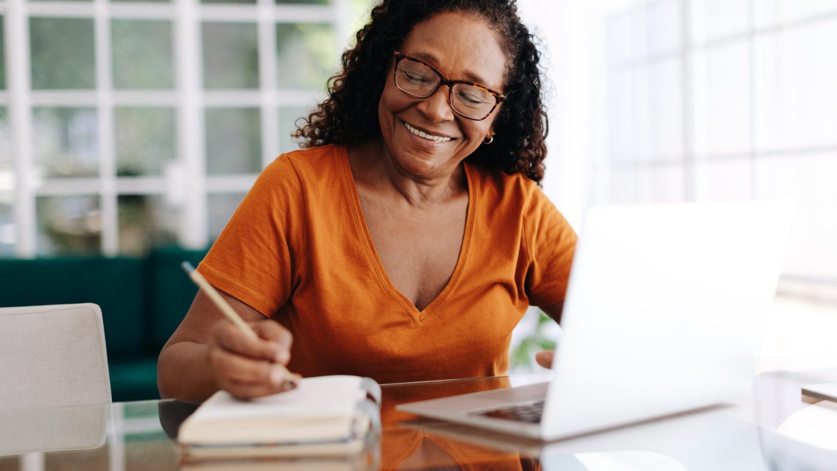 Happy woman sitting at a table in her home office using laptop and writing notes in a notepad